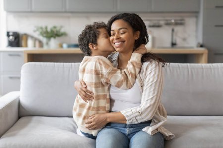 Foto de Concepto del Día de las Madres. Niño negro besando a su mamá en la mejilla, mamá afroamericana feliz e hijo preadolescente uniéndose en casa, disfrutando de momentos tiernos, relajándose en el sofá en la sala de estar - Imagen libre de derechos