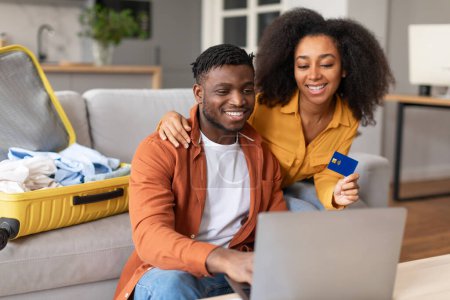 Photo for Happy african american tourists couple using laptop and credit card booking tour online, sitting with packed suitcase at modern home interior, spouses paying for hotel room reservation - Royalty Free Image