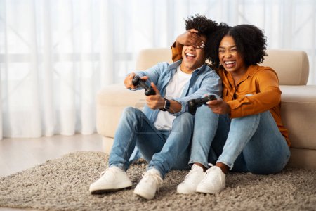 Téléchargez les photos : Une femme afro-américaine heureuse couvre ludique les yeux d'un homme pendant qu'ils rient et tiennent des contrôleurs de jeu, profitant d'une séance de jeu amusante ensemble sur le sol à la maison - en image libre de droit