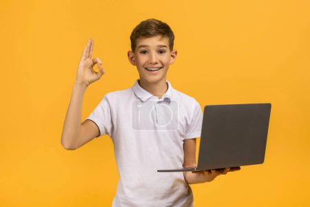 Foto de Niño adolescente confiado sosteniendo el ordenador portátil y haciendo el gesto de la mano bien, niño adolescente alegre usando polo blanco de pie aislado sobre el fondo amarillo, disfrutando de las tecnologías modernas - Imagen libre de derechos