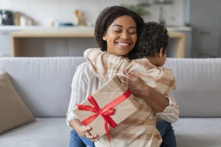 Foto de Feliz madre negra sosteniendo regalo envuelto con cinta roja y abrazando a su hijo, familia afroamericana de dos momentos de afecto y gratitud en su hogar, primer plano - Imagen libre de derechos