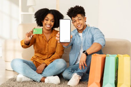 Téléchargez les photos : Un couple souriant est assis sur le sol, montrant un smartphone avec un écran vierge et une carte de crédit, indiquant un achat en ligne réussi, avec des sacs à provisions colorés à côté d'eux - en image libre de droit