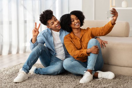 Photo for Sitting comfortably on the floor, a delighted young african american couple enjoys a light-hearted moment taking a selfie, with the man giving a peace sign and both wearing stylish denim - Royalty Free Image