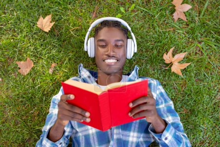 Photo for Contented young black man lies on grass engrossed in a book, headphones suggesting audiobook accompaniment to his reading, above view shot of guy enjoying novel outside - Royalty Free Image