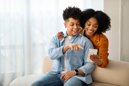 Téléchargez les photos : Un jeune homme afro-américain vêtu d'une chemise en denim et une femme vêtue d'un haut en moutarde sont assis sur un canapé, souriants et engagés avec un smartphone, rayonnant de chaleur et de convivialité - en image libre de droit
