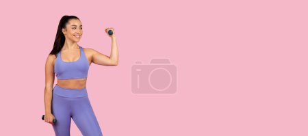 Téléchargez les photos : Femme en forme souriante dans l'ensemble d'entraînement de lavande cohésif effectue avec confiance biceps boucle avec haltères sur fond rose pastel, panorama, espace libre - en image libre de droit