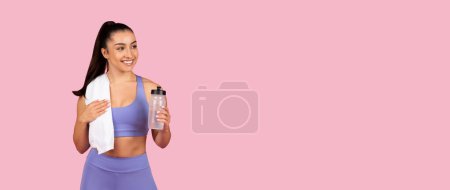 Foto de Mujer joven y renovada en sujetador deportivo púrpura y polainas sosteniendo botella de agua y toalla blanca sobre su hombro después de la sesión de gimnasio sobre fondo rosa, panorama, espacio libre - Imagen libre de derechos