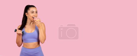 Téléchargez les photos : Femme de fitness joyeuse en vêtements de sport lavande aime beignet rose saupoudré tout en tenant haltère, mélange d'entraînement et de collation sur fond rose, panorama, espace libre - en image libre de droit