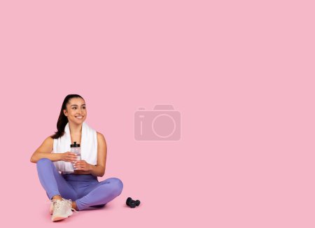 Foto de Mujer sonriente en traje de yoga sentada con botella de agua y toalla, tomando un descanso después de la rutina de fitness, con mancuerna cerca sobre fondo rosa, espacio libre - Imagen libre de derechos