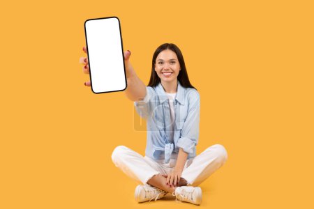 Téléchargez les photos : Joyeux jeune femme en tenue décontractée assise jambes croisées sur fond jaune, affichant joyeusement l'écran vierge du smartphone à la caméra, maquette - en image libre de droit