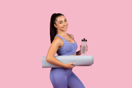 Foto de Radiante entusiasta del fitness en traje de yoga lavanda sosteniendo botella de agua y esterilla de yoga, preparándose para la rutina de bienestar en el fondo rosa - Imagen libre de derechos