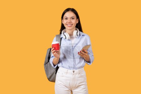 Photo for Smiling young woman with headphones around her neck holding red coffee cup and smartphone, wearing backpack on yellow background - Royalty Free Image