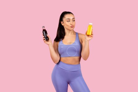 Photo for Athletic woman in sportswear holds soda and fresh juice bottle, contemplating choices, standing on vibrant pink background - Royalty Free Image