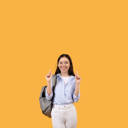 Téléchargez les photos : Jeune étudiante souriante pointant vers le haut à l'espace libre avec les deux mains, portant un sac à dos gris et souriant sur fond jaune vif, bannière - en image libre de droit