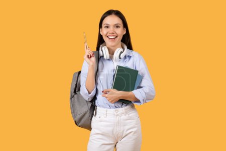 Téléchargez les photos : Joyeux jeune étudiante portant un sac à dos et des livres verts, portant des écouteurs blancs autour de son cou, tenant un stylo et souriant sur fond jaune - en image libre de droit