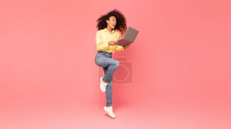 Foto de Mujer negra excitada saltando con emoción mientras sostiene el portátil, su alegría irradiando sobre un vibrante fondo rosa, ilustrando el entusiasmo y el éxito de la tecnología - Imagen libre de derechos