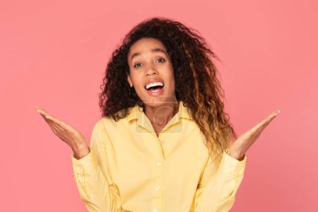 Photo for Amazed black woman in yellow blouse with hands raised in questioning gesture, mouth open in surprise, against plain pink background - Royalty Free Image