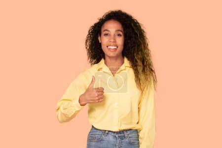 Téléchargez les photos : Femme noire confiante aux cheveux bouclés, les pouces souriants et gestuels levés, vêtus d'un chemisier jaune, sur un fond doux couleur pêche - en image libre de droit