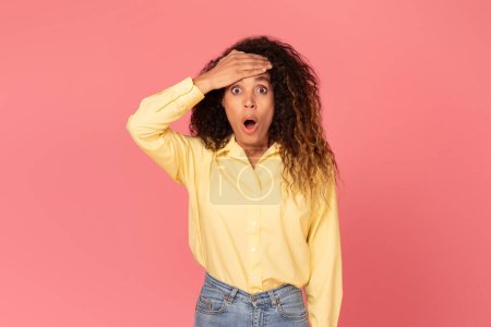 Photo for Surprised black woman, hand on her forehead and wide-eyed, gazes at camera with open mouth in shock against vivid pink background, lady conveying astonishment and unexpected reactions - Royalty Free Image