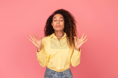 Photo for Peaceful black woman in yellow shirt with eyes closed and fingers in mudra pose, practicing meditation against tranquil pink backdrop - Royalty Free Image