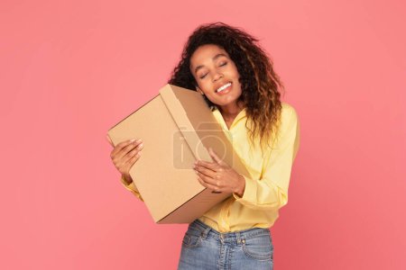 Photo for Cheerful, young black woman smiling as embraces box parcel, radiating happiness against vibrant pink background, expressing excitement, receiving gifts, and joyful moments - Royalty Free Image