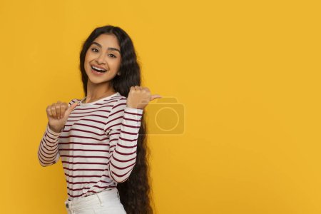 Photo for Great offer, exciting deal. Smiling attractive long-haired young indian woman pointing at blank copy space mockup for advertisement isolated on yellow studio background - Royalty Free Image