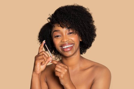 Téléchargez les photos : Femme noire lumineuse et joyeuse appliquant le maquillage au pinceau, son sourire exprimant le plaisir, placé sur un fond beige chaud qui accentue sa beauté - en image libre de droit