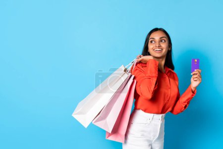 Photo for Content hindu customer lady holding credit card and paper shopper bags advertising shopping offer, looking at free space for text on blue studio backdrop, commerce and consumerism - Royalty Free Image