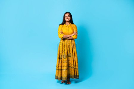 Photo for Confident Indian Woman. Smiling young hindu lady wearing yellow dress posing with folded arms standing isolated on blue studio backdrop. Happy casual person, full length shot - Royalty Free Image