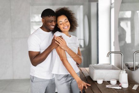 Photo for Loving Black Husband Hugging His Happy Beautiful Wife Standing In Modern Bathroom Together, Enjoying Romance Of Daily Beauty Routine In The Morning. Family Pampering And Self Care - Royalty Free Image