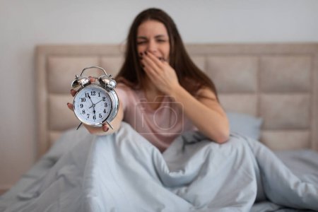 Photo for Sleepy young woman yawns as she sits in bed, presenting classic alarm clock to the camera, illustrating the struggle of an early wake-up, in the morning - Royalty Free Image