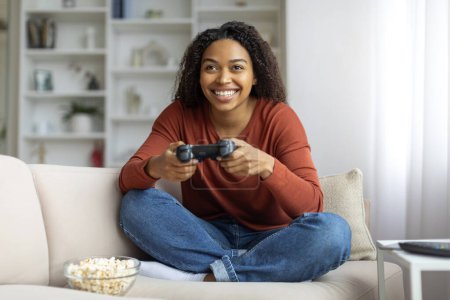 Foto de Diversión doméstica. Joven y alegre mujer negra jugando videojuegos en casa, feliz dama afroamericana usando joystick y sonriendo mientras está sentada en el sofá con palomitas de maíz en la sala de estar, espacio libre - Imagen libre de derechos