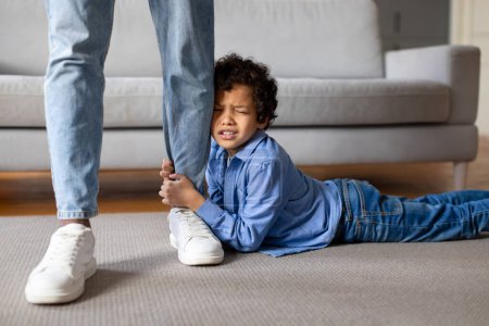 Photo for Distressed black boy holding onto his fathers leg tightly, showing plea for attention or refusal to let go, while the parent stands firm, home interior - Royalty Free Image