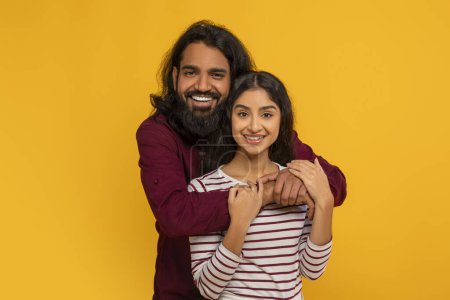 Téléchargez les photos : Portrait de joyeux amoureux jeune couple hindou posant sur fond de studio jaune. Joyeux beau mec indien étreignant par derrière sa jolie petite amie. Amour, relations, affection - en image libre de droit