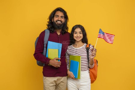 Foto de Feliz joven indio hombre y mujer llevando mochilas y libros de ejercicios posando sobre fondo de estudio amarillo, estudiantes orientales amigos mostrando la bandera de EE.UU.. Educación en el extranjero, aprendizaje de lenguas extranjeras - Imagen libre de derechos