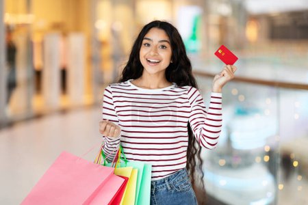 Photo for Black friday, season sale. Excited young indian woman enjoying shopping at newest modern mall, carrying colorful paper bags purchases and showing credit card. Contactless shopping - Royalty Free Image