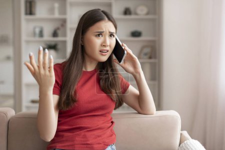Photo for Perplexed young asian woman gesturing with her hand while talking on the phone at home, korean lady expressing confusion, sitting on beige couch in light-filled, modern living room, copy space - Royalty Free Image