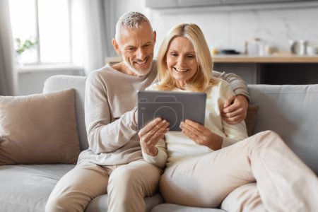 Téléchargez les photos : Couples retraités séduisants positifs utilisant une tablette numérique à la maison, allongés sur le canapé dans un studio confortable, embrassant et regardant l'écran d'ordinateur, magasinant sur les services bancaires en ligne, lisant le blog - en image libre de droit