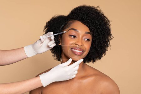 Photo for Smiling young black woman receiving professional cosmetic treatment with syringe, demonstrating beauty procedure against beige background - Royalty Free Image