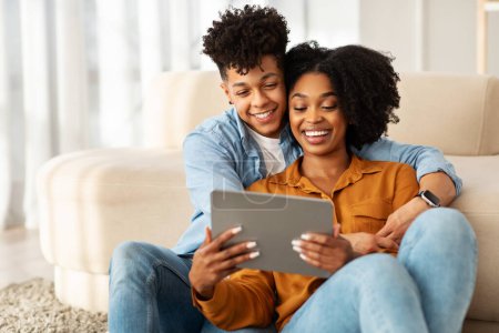 Foto de Acurrucado en el sofá, la joven pareja afroamericana en ropa casual comparte un momento tierno mientras miran felizmente una tableta juntos en una acogedora y luminosa sala de estar - Imagen libre de derechos