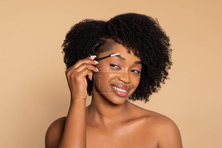 Téléchargez les photos : Jeune femme noire avec un sourire radieux toilettant ses sourcils avec une brosse blanche, mettant en valeur la routine quotidienne de beauté sur fond beige doux - en image libre de droit