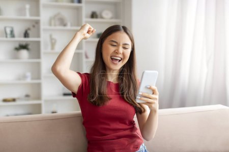 Photo for Euphoric young asian woman cheering success with smartphone at home, happy korean lady emotionally reacting to pleasant message, got good news, sitting on couch in well-lit living room, copy space - Royalty Free Image
