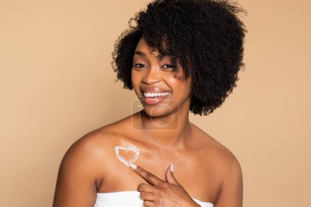Téléchargez les photos : Joyeuse femme noire enveloppée dans une serviette pointant vers une lotion en forme de cœur sur son épaule, symbolisant l'amour pour les soins de la peau sur fond beige doux - en image libre de droit