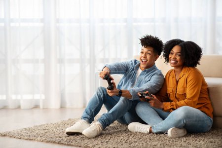 Foto de Una alegre pareja joven afroamericana disfruta de un momento lúdico juntos mientras se sientan en el suelo y participan en una competencia de videojuegos en una sala de estar luminosa y acogedora - Imagen libre de derechos