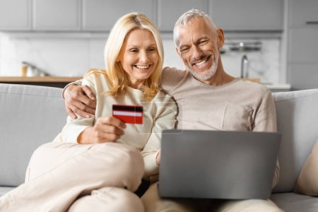 Photo for Smiling attractive caucasian retired man and woman shopping online together, using laptop computer pc and red plastic bank credit card, sitting on couch at home. Retail, ecommerce - Royalty Free Image