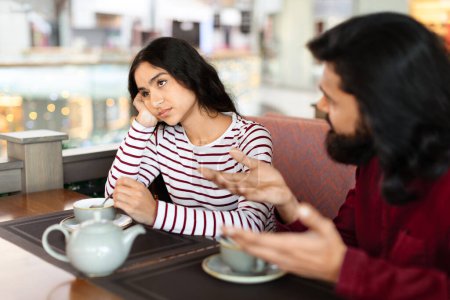 Photo for Indian couple have fight at cafe at shopping mall, drinking tea. Young pretty eastern lady feeling bored while her boyfriend complaining or explaining something, copy space - Royalty Free Image