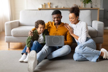 Téléchargez les photos : Famille noire souriante assise sur le sol avec un ordinateur portable, partageant un moment joyeux alors qu'ils s'engagent dans une activité en ligne ensemble dans leur maison, surfant sur Internet sur ordinateur - en image libre de droit