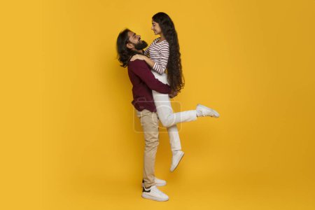Photo for Loving handsome bearded young indian guy lifting up his beautiful long-haired girlfriend, cheerful romantic hindu couple enjoying time together isolated on yellow background, copy space, full length - Royalty Free Image