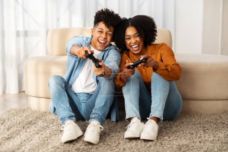 Téléchargez les photos : Un heureux couple afro-américain assis sur le sol avec des contrôleurs de jeu à la main, profondément engagé et s'éclater tout en jouant à des jeux vidéo dans un salon lumineux - en image libre de droit
