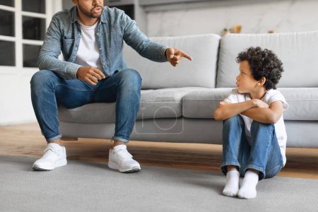Téléchargez les photos : Jeune père noir gronder son fils préadolescent à la maison, pointant du doigt dans un geste disciplinaire à son enfant mâle contrarié assis sur le sol avec les bras croisés dans le salon minimaliste, recadré - en image libre de droit